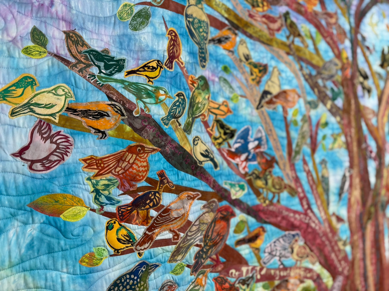 A close-up of a colorful quilted textile featuring many birds perched on tree branches against a blue sky-like background.