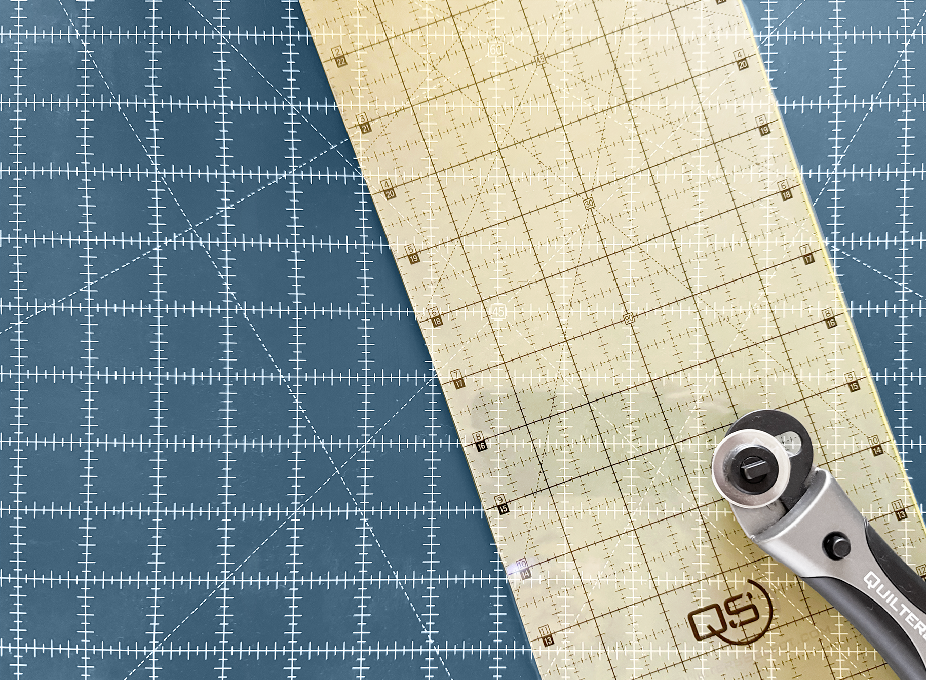 A clear acrylic quilting ruler with a beige grid overlay, shown diagonally on a blue cutting mat with a rotary cutter in the bottom right corner.