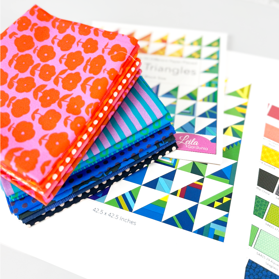 A stack of colorful fabric quilts with floral and striped patterns, arranged on top of printed quilting guides and a color chart. A pink tag reads 'Lula Quintaro'.